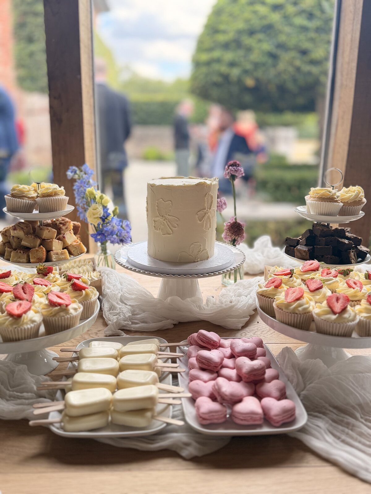 Wedding dessert table with a buttercream cutting cake surrounded by cupcakes, brownies, blondies, cakesicles and heart macarons