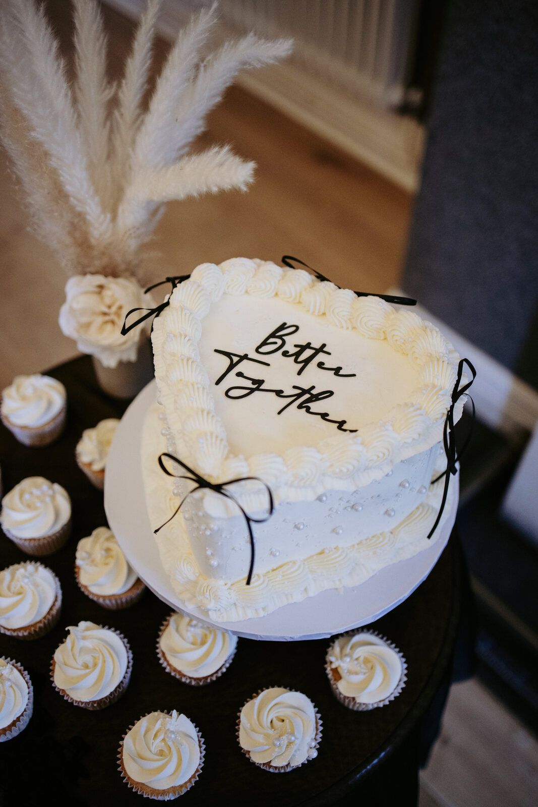 Heart-shaped Lambeth wedding cake with black ribbon bows and a 'Better Together' topper, surrounded by cupcakes