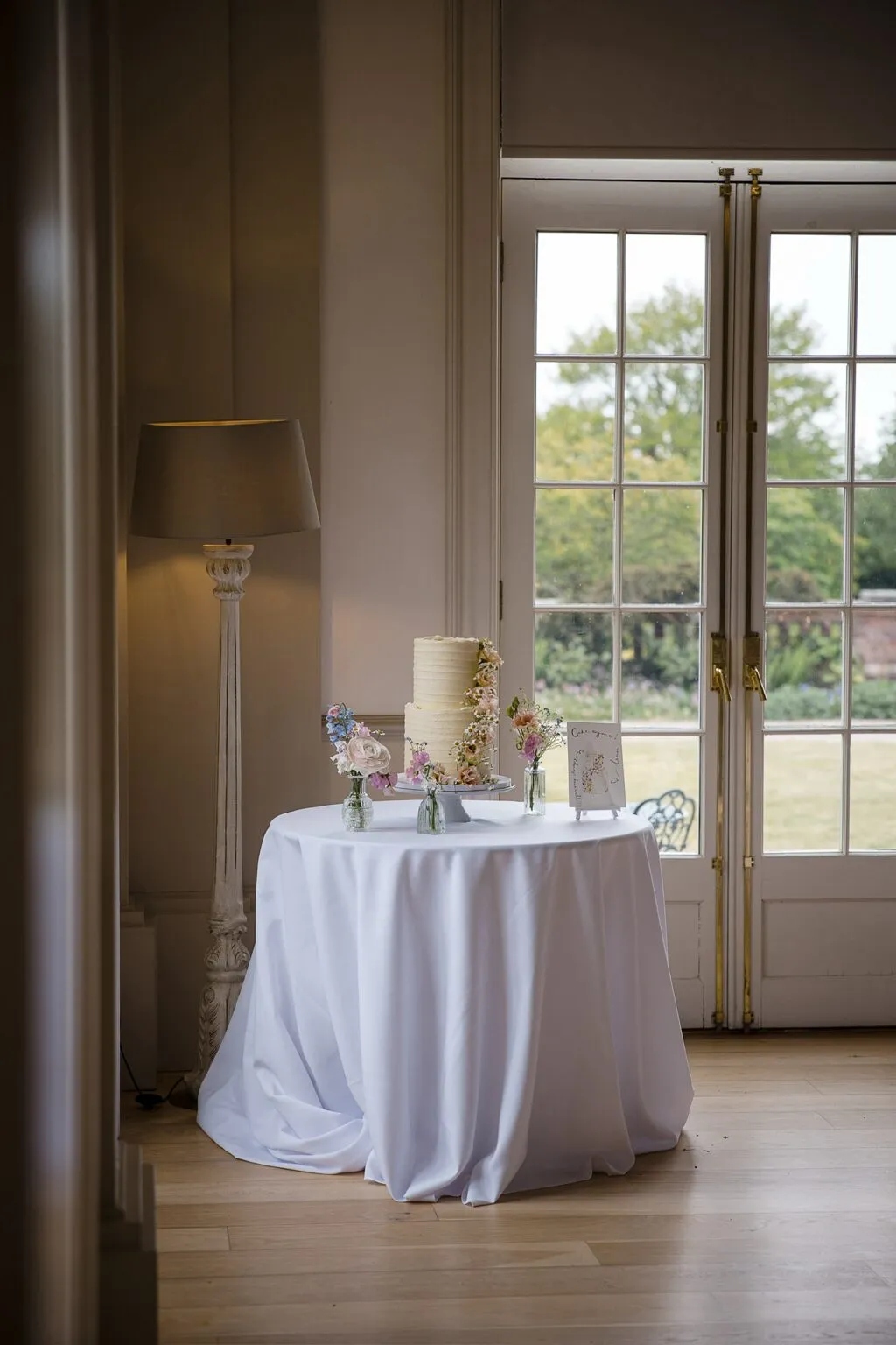Soft buttercream wedding cake with a natural floral cascade beside the tall windows at Hodsock Priory