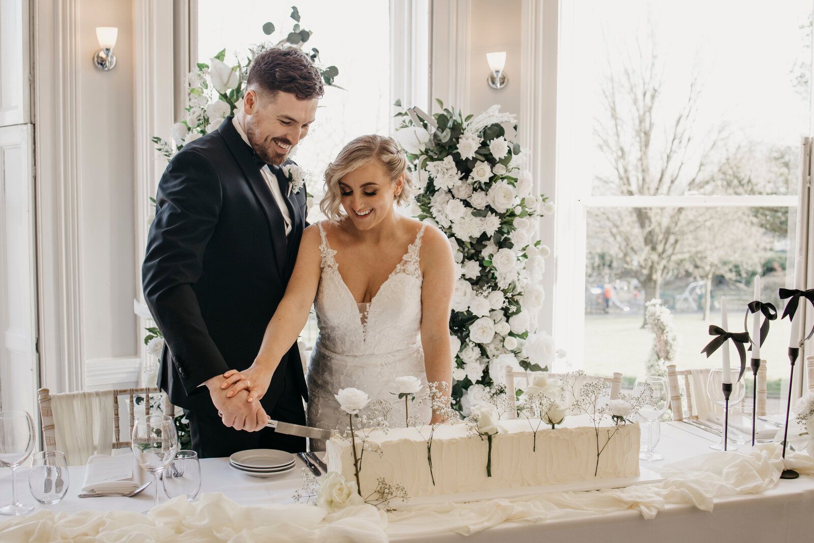 Bride and groom cutting a long buttercream wedding cake decorated with white flowers