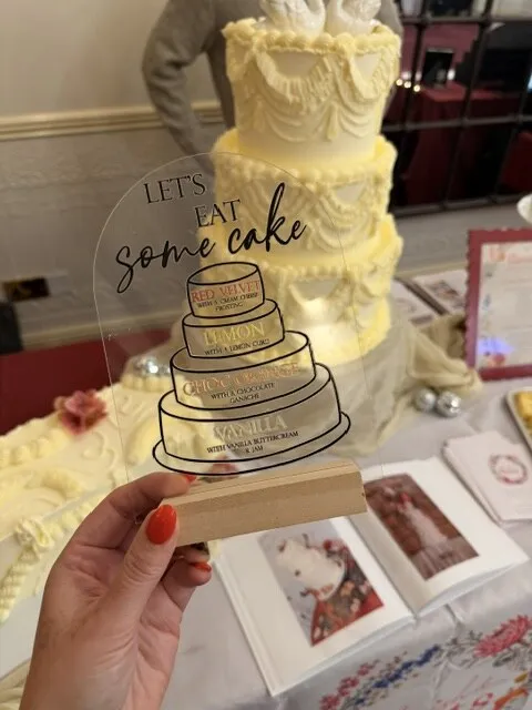 A ‘Let’s eat some cake’ flavour sign held up in front of a Lambeth-piped wedding cake