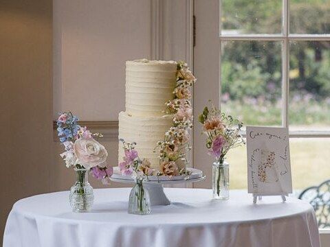Two tier textured buttercream wedding cake with a cascade of pastel flowers, beside bud vases and an illustrated cake menu